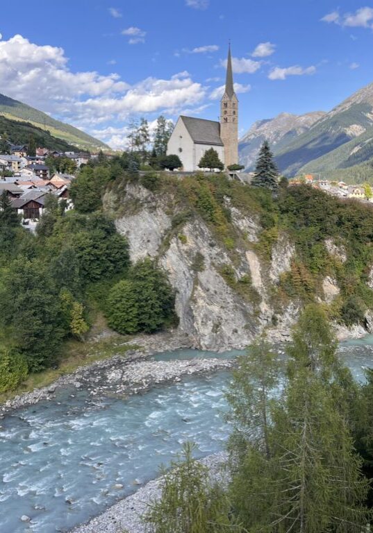 Idyllisches Bergdorf mit Kirche am Flussufer, möglicherweise Teil des Transalpine Run 2025. - We Run 4 Fun!